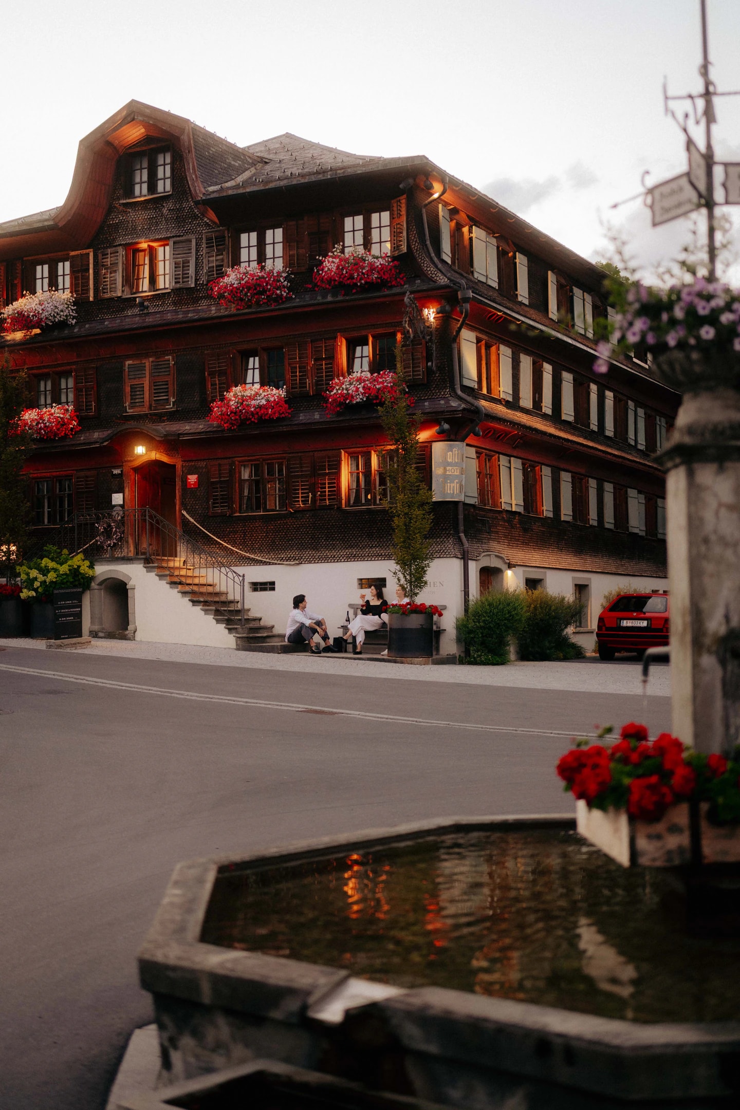 Der Hirschen am Dorfplatz im Sommer, Geranien an allen Fenstern, vorne der Dorfbrunnen.